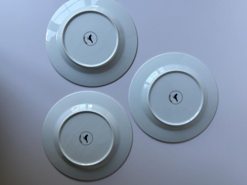 May include: Three white ceramic plates, viewed from the underside, arranged on a light surface. Each plate has a circular indentation in the center with a black logo. The plates are clean and appear unused.