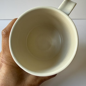 May include: Cream-colored ceramic mug with a unique handle design. The mug's interior is a soft, off-white hue, and the handle is a rectangular shape that curves slightly. The mug is held in a hand, showcasing its size and form.