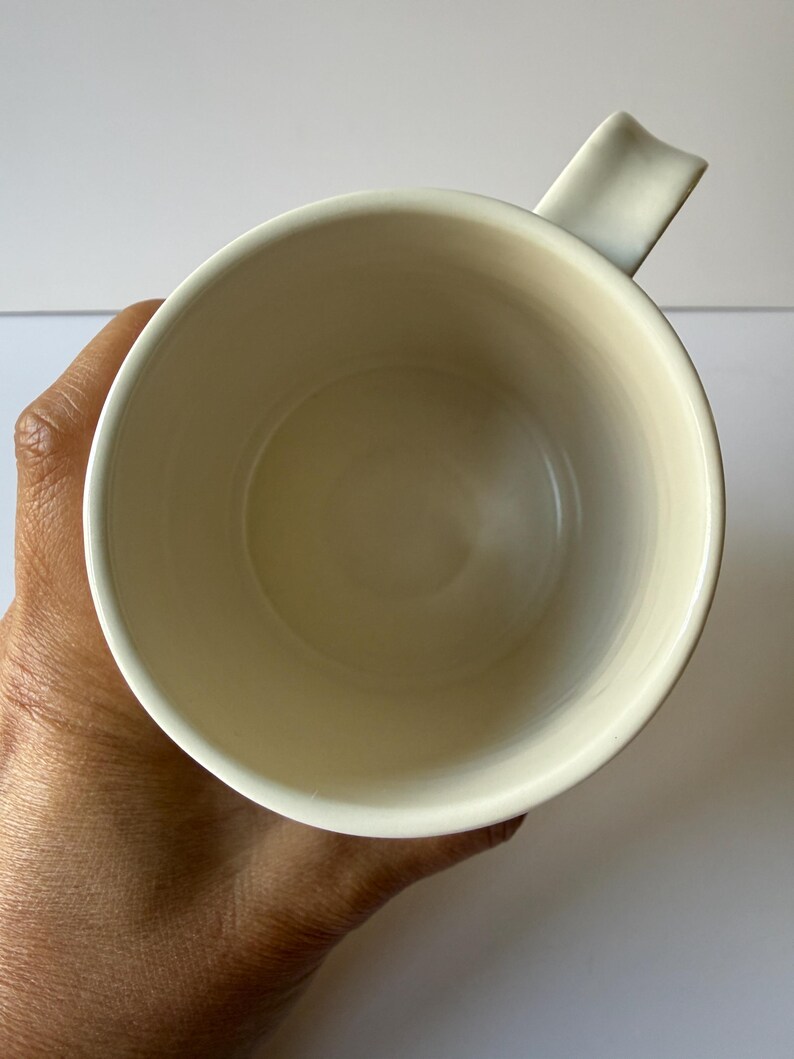 May include: Cream-colored ceramic mug with a unique handle design. The mug's interior is a soft, off-white hue, and the handle is a rectangular shape that curves slightly. The mug is held in a hand, showcasing its size and form.