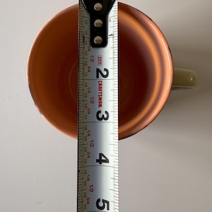 Puede incluir: Vista superior de una taza de color cobre con un asa verde claro. Una cinta m&eacute;trica dentro de la taza indica una medida de aproximadamente 12,7 cm. El interior de la taza tiene un acabado brillante.