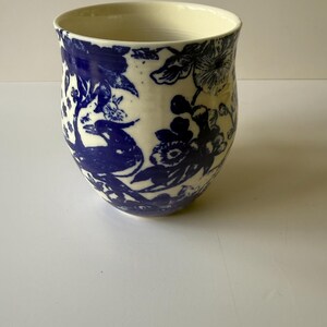 May include: A ceramic vase with a cream-colored interior and a white exterior. The exterior is decorated with a blue floral pattern. The vase has a rounded shape and a slightly flared rim. The design features flowers and foliage.