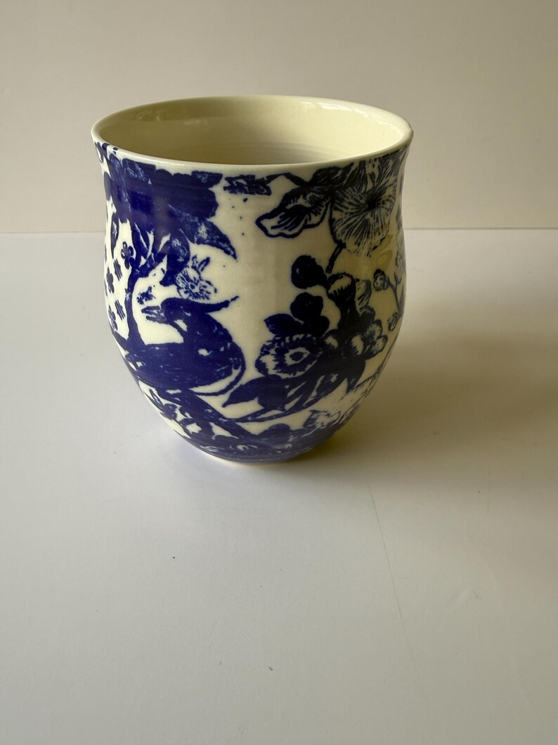 May include: A ceramic vase with a cream-colored interior and a white exterior. The exterior is decorated with a blue floral pattern. The vase has a rounded shape and a slightly flared rim. The design features flowers and foliage.