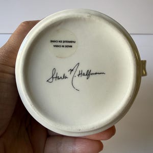 May include: Cream-colored ceramic mug base with a gold rim, held by a hand. The bottom of the mug features a sticker with text that reads "MADE IN CHINA FABRIQUE EN CHINE" and a signature. The mug is likely a collectible or decorative item.