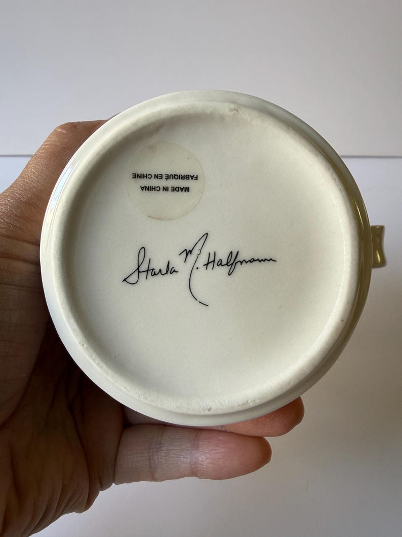 May include: Cream-colored ceramic mug base with a gold rim, held by a hand. The bottom of the mug features a sticker with text that reads "MADE IN CHINA FABRIQUE EN CHINE" and a signature. The mug is likely a collectible or decorative item.