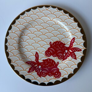 May include: Round decorative plate with a gold rim and a scalloped edge. The plate features a repeating wave pattern in cream and orange, with two red goldfish designs. A stylish addition to any home decor.