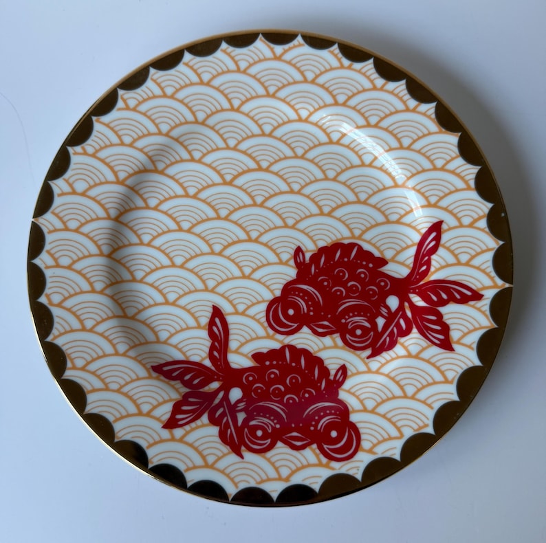 May include: Round decorative plate with a gold rim and a scalloped edge. The plate features a repeating wave pattern in cream and orange, with two red goldfish designs. A stylish addition to any home decor.
