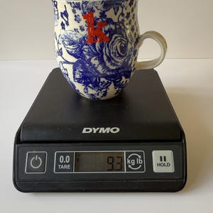 May include: A ceramic mug with a blue floral pattern and a red letter "K" is on a black scale. The mug has a white interior and a curved handle. The scale displays a weight of 9.3 lb.