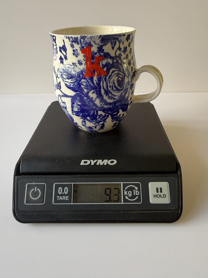 May include: A ceramic mug with a blue floral pattern and a red letter "K" is on a black scale. The mug has a white interior and a curved handle. The scale displays a weight of 9.3 lb.