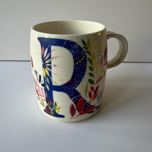May include: Cream-colored ceramic mug featuring a large, blue letter "R" adorned with floral patterns in red, yellow, and pink. The mug has a curved handle and is decorated with colorful leaves and flowers.