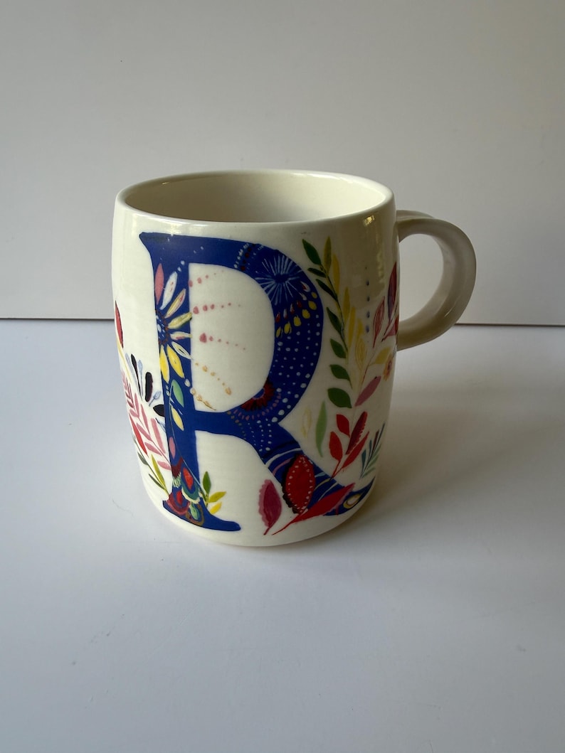 May include: Cream-colored ceramic mug featuring a large, blue letter "R" adorned with floral patterns in red, yellow, and pink. The mug has a curved handle and is decorated with colorful leaves and flowers.