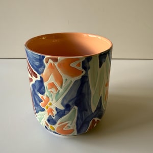 Puede incluir: Una taza de cer&aacute;mica con un dise&ntilde;o floral. El exterior presenta un fondo azul oscuro con patrones florales naranjas, blancos y rojos. El interior es de color melocot&oacute;n.