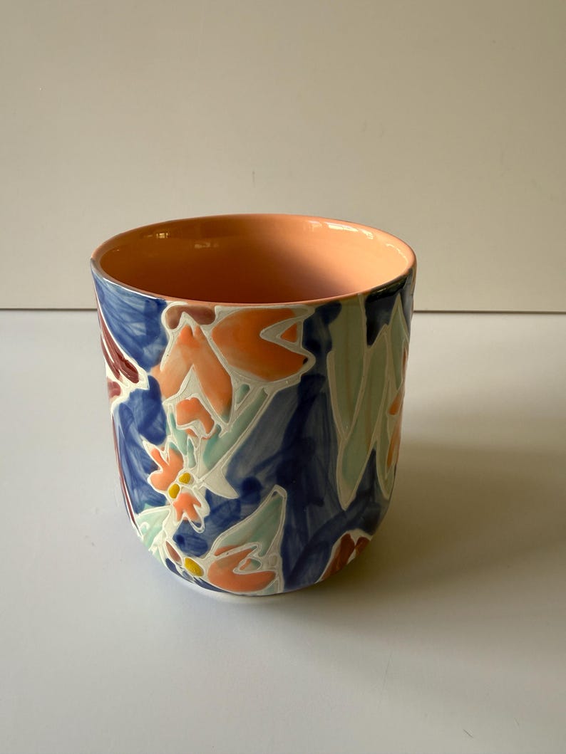 Puede incluir: Una taza de cer&aacute;mica con un dise&ntilde;o floral. El exterior presenta un fondo azul oscuro con patrones florales naranjas, blancos y rojos. El interior es de color melocot&oacute;n.