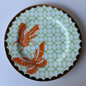 May include: A decorative plate featuring two orange butterfly designs. The plate has a light green and white patterned background with a scalloped brown border and a gold rim. The plate is round.