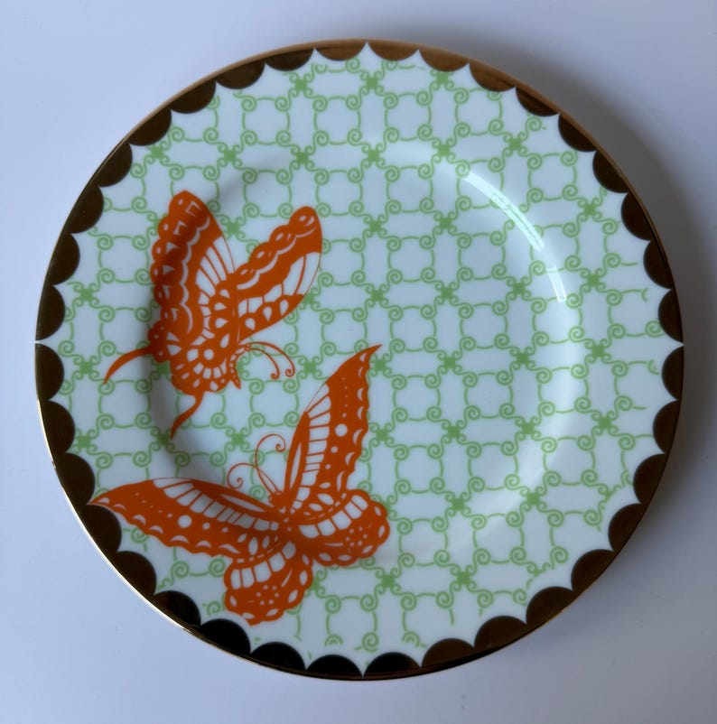 May include: A decorative plate featuring two orange butterfly designs. The plate has a light green and white patterned background with a scalloped brown border and a gold rim. The plate is round.