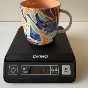 Puede incluir: Una taza de cer&aacute;mica con un dise&ntilde;o floral en tonos azules, naranjas y verdes. El interior es color melocot&oacute;n y el asa es gris. La taza est&aacute; sobre una b&aacute;scula digital negra, que marca 5,3 kg.