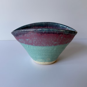 May include: A handcrafted ceramic bowl with a distinctive form and colour palette. It has a dark blue rim, a band of reddish-purple, and a turquoise-green lower section. The bowl has a speckled texture and a cream-coloured base.