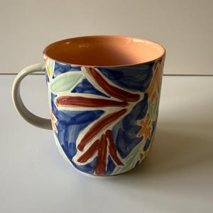 Puede incluir: Una taza de cer&aacute;mica con un dise&ntilde;o floral. La taza presenta un fondo azul con patrones florales rojos, naranjas y verdes. El interior es de color melocot&oacute;n y el asa es gris.