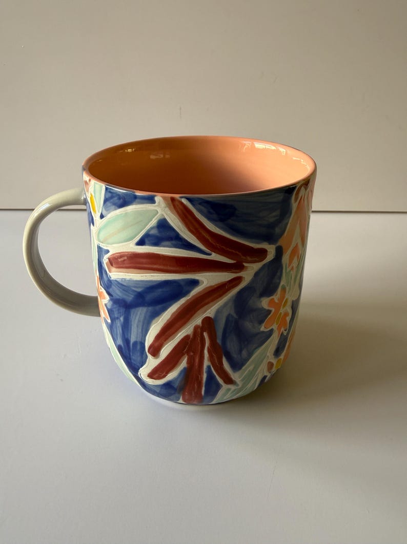 Puede incluir: Una taza de cer&aacute;mica con un dise&ntilde;o floral. La taza presenta un fondo azul con patrones florales rojos, naranjas y verdes. El interior es de color melocot&oacute;n y el asa es gris.