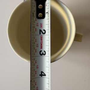 May include: A cream-colored ceramic mug with a handle, measured with a Craftsman tape measure. The mug is approximately 4 inches tall. The interior of the mug is visible, and the handle is on the right side.