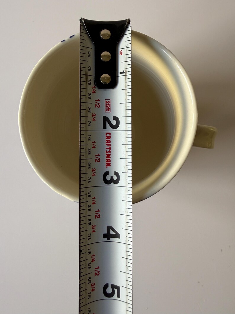 May include: A cream-colored ceramic mug with a handle, measured with a Craftsman tape measure. The mug is approximately 4 inches tall. The interior of the mug is visible, and the handle is on the right side.