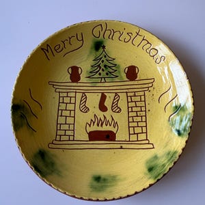 May include: A round yellow ceramic plate with a hand-painted brown illustration of a fireplace featuring a Christmas tree, two stockings, and two pitchers. The text Merry Christmas is written in brown at the top, with green accents on the rim.