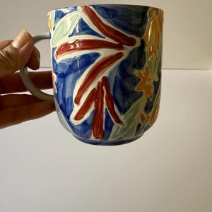 Puede incluir: Una taza de cer&aacute;mica con un asa gris y un dise&ntilde;o floral vibrante. La taza presenta un fondo azul oscuro con flores y hojas pintadas en rojo, blanco, amarillo y verde. La taza est&aacute; sujeta por una mano.