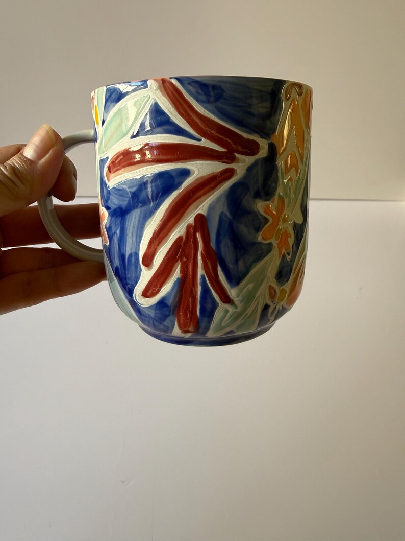 Puede incluir: Una taza de cer&aacute;mica con un asa gris y un dise&ntilde;o floral vibrante. La taza presenta un fondo azul oscuro con flores y hojas pintadas en rojo, blanco, amarillo y verde. La taza est&aacute; sujeta por una mano.