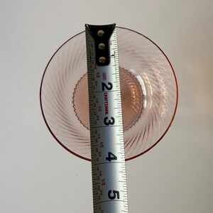 May include: A pink glass plate with a spiral design, approximately 5 inches in diameter, is measured with a Craftsman tape measure. The plate has a raised rim and a textured center. The tape measure shows the plate's diameter.