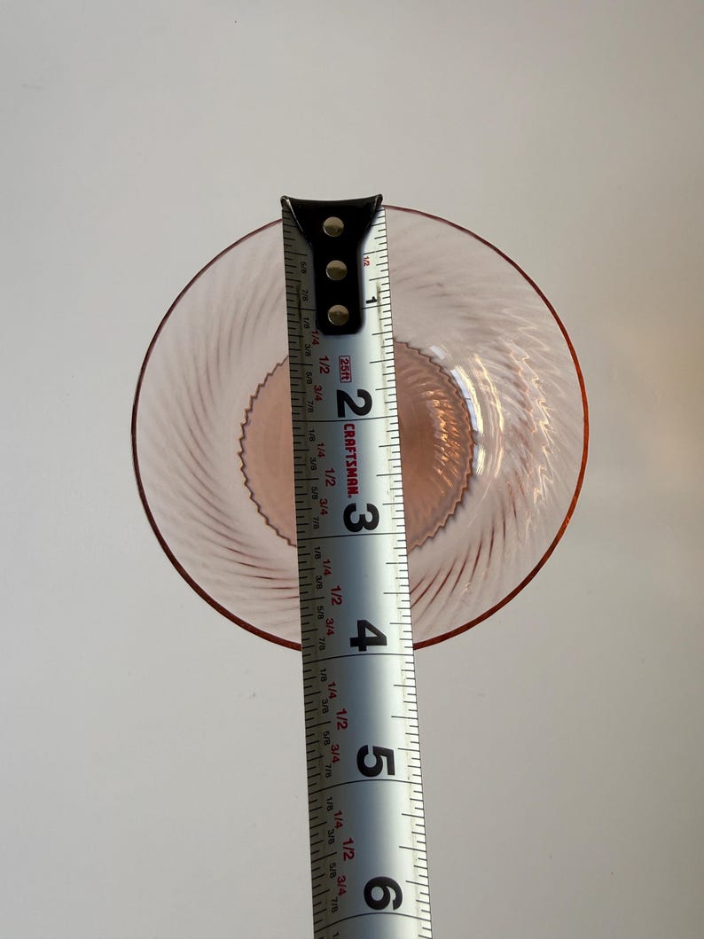 May include: A pink glass plate with a spiral design, approximately 5 inches in diameter, is measured with a Craftsman tape measure. The plate has a raised rim and a textured center. The tape measure shows the plate's diameter.