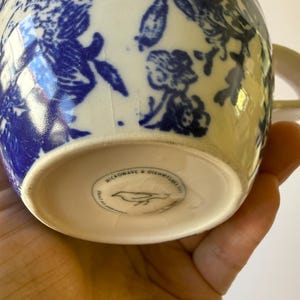 May include: A ceramic mug with a blue floral pattern on a white background. The mug has a handle and the bottom of the mug has the text "MICROWAVE & DISHWASHER SAFE" and a bird logo.