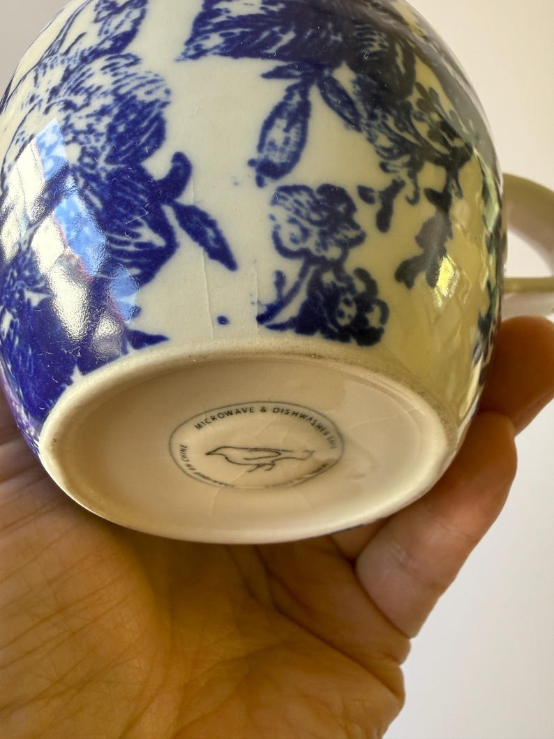 May include: A ceramic mug with a blue floral pattern on a white background. The mug has a handle and the bottom of the mug has the text "MICROWAVE & DISHWASHER SAFE" and a bird logo.