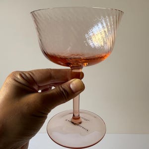 May include: A pink-tinted glass with a textured bowl and a slender stem. The glass has a wide, shallow bowl with a swirling pattern. The base of the glass is round. The text "Anthropologie" is printed on the base.