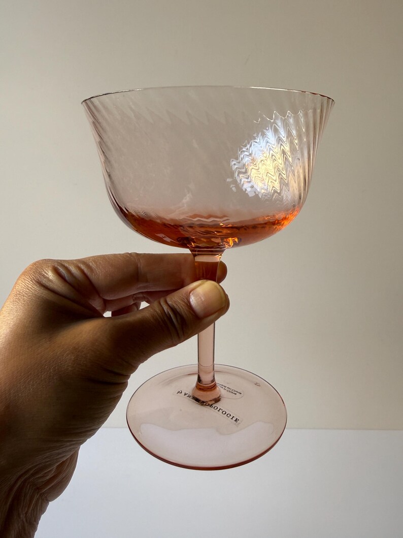 May include: A pink-tinted glass with a textured bowl and a slender stem. The glass has a wide, shallow bowl with a swirling pattern. The base of the glass is round. The text "Anthropologie" is printed on the base.