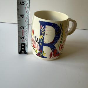 May include: Cream-colored ceramic mug with a large blue letter "R" decorated with colorful floral patterns. The mug is approximately 4 inches tall and has a handle on the right side. The mug is on a white surface.