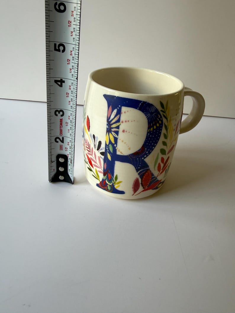 May include: Cream-colored ceramic mug with a large blue letter "R" decorated with colorful floral patterns. The mug is approximately 4 inches tall and has a handle on the right side. The mug is on a white surface.
