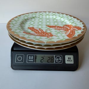 May include: A stack of three decorative plates with a light green and gold rim design. Each plate features a repeating pattern and two orange butterfly illustrations. The plates are on a black digital scale, weighing 2.8 lbs.