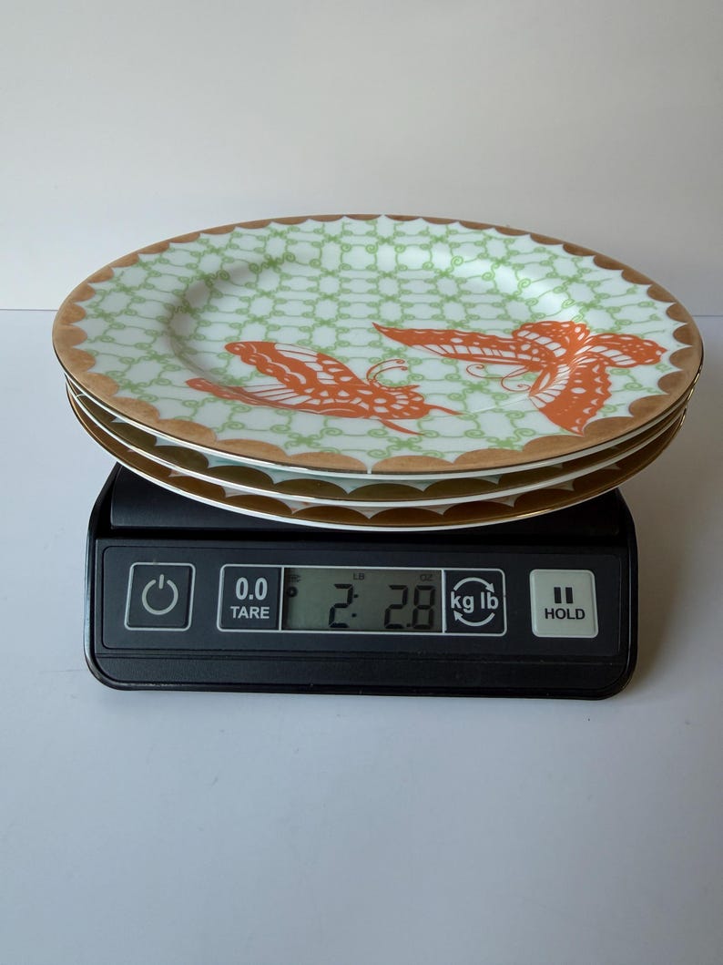 May include: A stack of three decorative plates with a light green and gold rim design. Each plate features a repeating pattern and two orange butterfly illustrations. The plates are on a black digital scale, weighing 2.8 lbs.