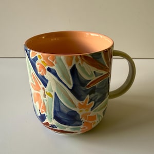 Puede incluir: Una taza de cer&aacute;mica con un dise&ntilde;o floral. El exterior presenta un patr&oacute;n de hojas y flores naranjas, azules y verdes. El interior es de color melocot&oacute;n y el asa es verde claro.