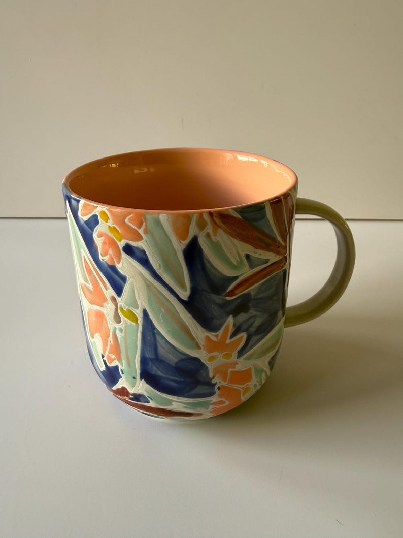 Puede incluir: Una taza de cer&aacute;mica con un dise&ntilde;o floral. El exterior presenta un patr&oacute;n de hojas y flores naranjas, azules y verdes. El interior es de color melocot&oacute;n y el asa es verde claro.