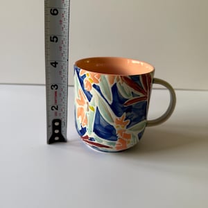 Puede incluir: Una taza de cer&aacute;mica con un dise&ntilde;o floral. La taza presenta un fondo azul con detalles florales en naranja, blanco y rojo. El interior es de color melocot&oacute;n y el asa es gris. La taza mide aproximadamente 10 cm de alto.
