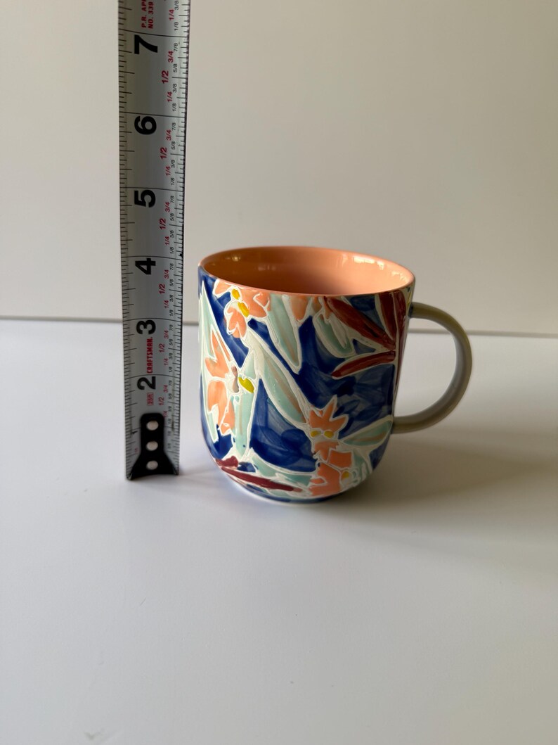 Puede incluir: Una taza de cer&aacute;mica con un dise&ntilde;o floral. La taza presenta un fondo azul con detalles florales en naranja, blanco y rojo. El interior es de color melocot&oacute;n y el asa es gris. La taza mide aproximadamente 10 cm de alto.