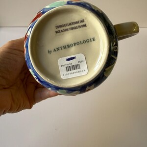 Puede incluir: Una taza de cer&aacute;mica con un dise&ntilde;o floral colorido. La taza tiene una base azul con detalles en rojo, verde y blanco. El asa es de color verde apagado. La parte inferior de la taza tiene el texto "by ANTHROPOLOGIE".