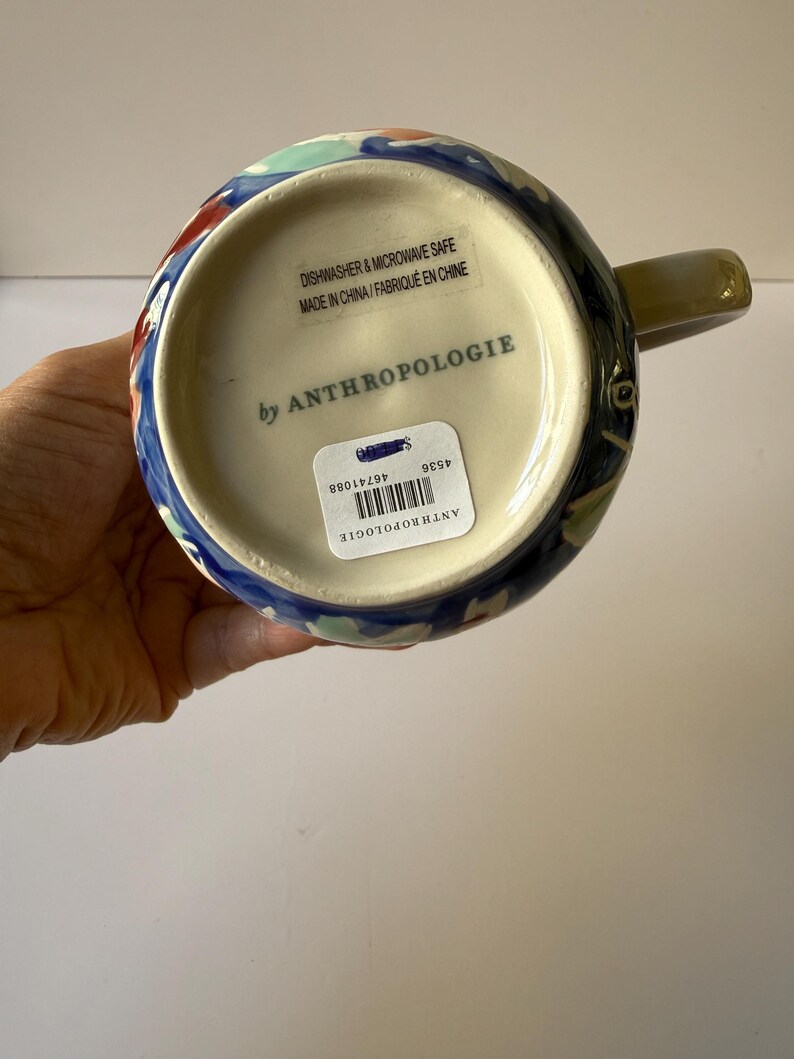 Puede incluir: Una taza de cer&aacute;mica con un dise&ntilde;o floral colorido. La taza tiene una base azul con detalles en rojo, verde y blanco. El asa es de color verde apagado. La parte inferior de la taza tiene el texto "by ANTHROPOLOGIE".