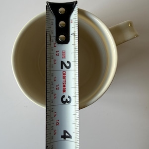 May include: A cream-colored mug with a handle, viewed from above. A measuring tape is placed next to the mug, showing a diameter of approximately 3 inches. The tape is marked with the Craftsman logo.