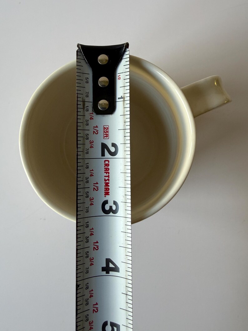 May include: A cream-colored mug with a handle, viewed from above. A measuring tape is placed next to the mug, showing a diameter of approximately 3 inches. The tape is marked with the Craftsman logo.