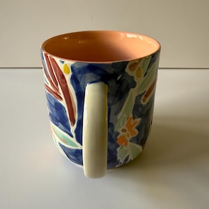 Puede incluir: Una taza de cer&aacute;mica con un asa blanca y un exterior azul decorado con coloridos estampados florales. El interior de la taza es de color melocot&oacute;n. La taza tiene forma cil&iacute;ndrica.