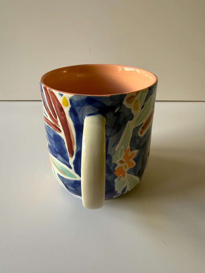 Puede incluir: Una taza de cer&aacute;mica con un asa blanca y un exterior azul decorado con coloridos estampados florales. El interior de la taza es de color melocot&oacute;n. La taza tiene forma cil&iacute;ndrica.