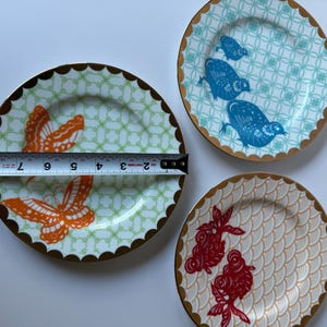 May include: Three decorative ceramic plates with gold scalloped edges. One plate features orange butterflies, another has blue birds, and the third has red fish. The plates measure approximately 8 inches in diameter.