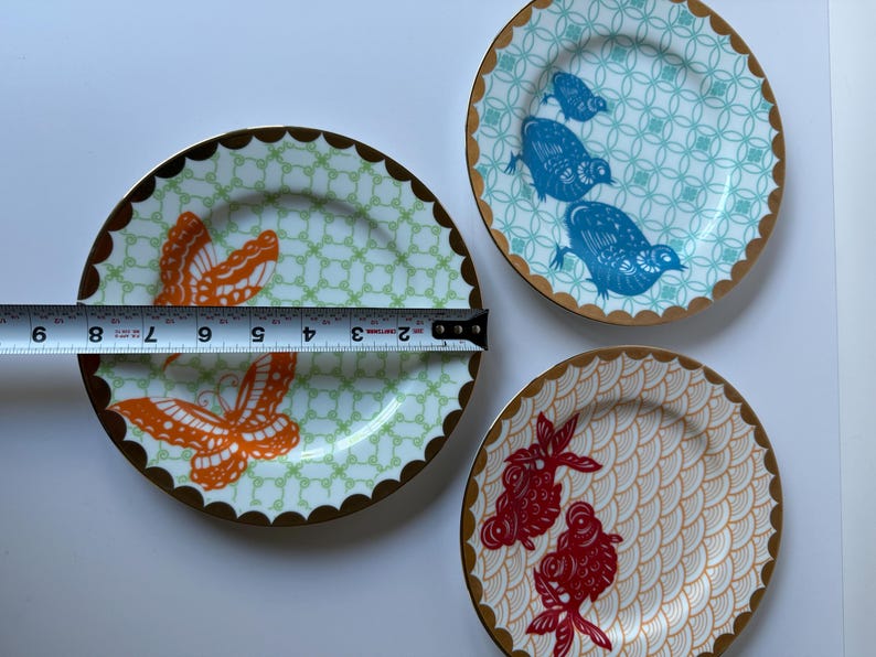 May include: Three decorative ceramic plates with gold scalloped edges. One plate features orange butterflies, another has blue birds, and the third has red fish. The plates measure approximately 8 inches in diameter.