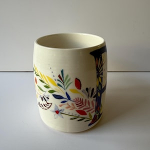 May include: A cream-colored ceramic mug with a floral design. The mug features a colorful illustration of leaves, flowers, and branches in shades of blue, yellow, red, and pink. The mug has a cylindrical shape and a smooth surface.